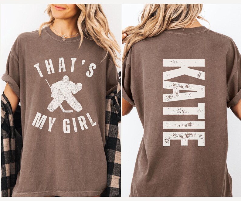 May include: A brown t-shirt with a white graphic of a hockey goalie and the text "That's My Girl". The back of the shirt has the name "Katie" in white text.