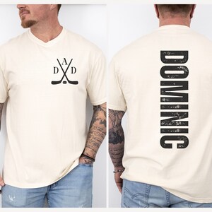 Puede incluir: Una camiseta blanca con un dise&ntilde;o de palo de hockey negro en la parte delantera y la palabra "DOMINIC" impresa verticalmente en la parte posterior.