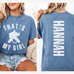 May include: A blue t-shirt with the text "That's My Girl" and a hockey goalie graphic on the front. The back of the shirt has the name "Hannah" printed vertically in white.