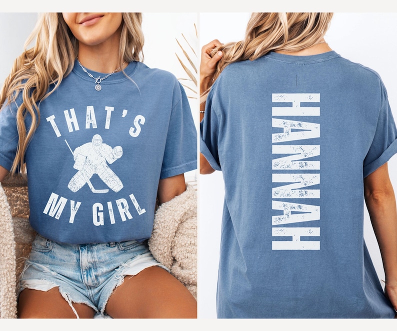 May include: A blue t-shirt with the text "That's My Girl" and a hockey goalie graphic on the front. The back of the shirt has the name "Hannah" printed vertically in white.