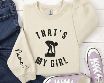 May include: Cream-colored sweatshirt with the text "THAT'S MY GIRL" and a swimmer graphic in black. The left sleeve has the word "Name" in cursive with a heart. A pair of jeans and purple sneakers are also in the image.