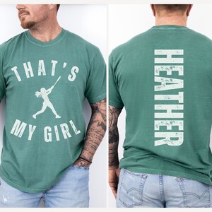 May include: A green t-shirt with a white graphic of a girl playing baseball and the text "That's My Girl". The back of the shirt has the name "Heather" printed vertically in white.