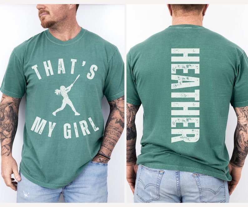 May include: A green t-shirt with a white graphic of a girl playing baseball and the text "That's My Girl". The back of the shirt has the name "Heather" printed vertically in white.