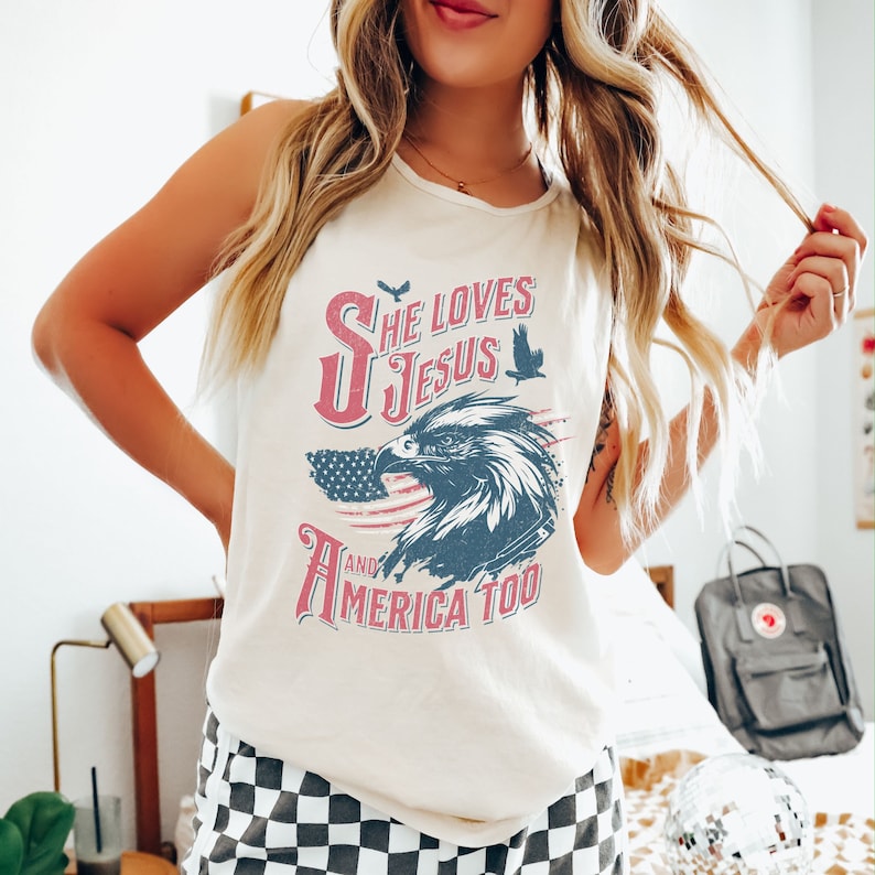 May include: White tank top with a red, white, and blue American flag design. The text "She Loves Jesus and America Too" is printed on the shirt with a vintage eagle graphic.