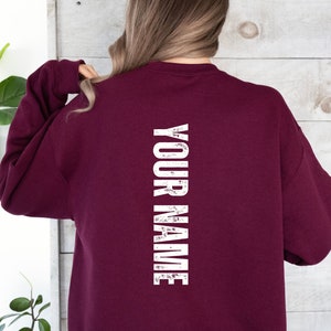 Może przedstawiać: Bordowa bluza z nadrukiem "YOUR NAME" w pionie na plecach, w kolorze białym, z efektem postarzenia.