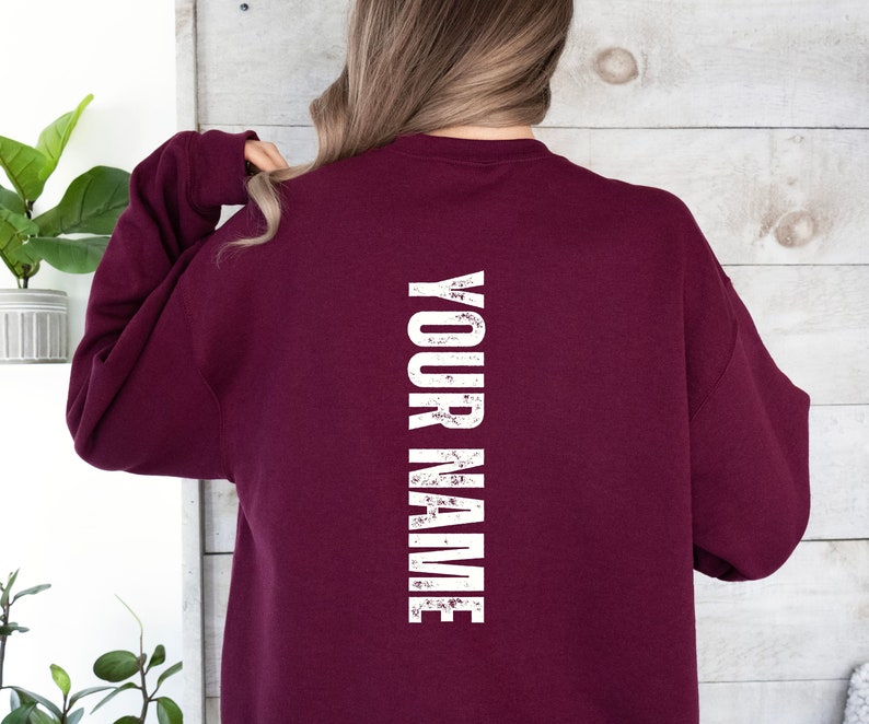 Może przedstawiać: Bordowa bluza z nadrukiem "YOUR NAME" w pionie na plecach, w kolorze białym, z efektem postarzenia.