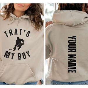 Może przedstawiać: Beżowa bluza z kapturem z czarną grafiką hokeisty i tekstem "That's My Boy" z przodu. Z tyłu bluzy z kapturem znajduje się tekst "Your Name" wydrukowany pionowo.