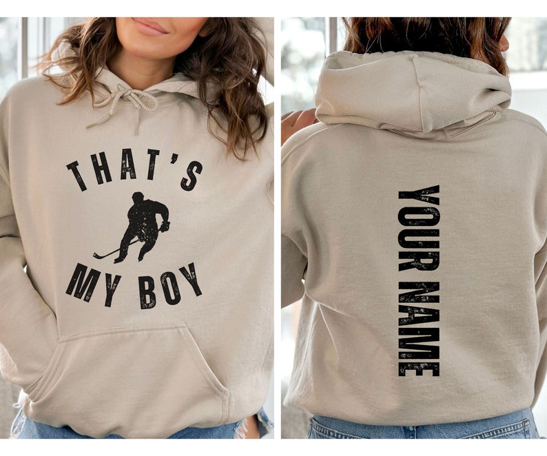 Może przedstawiać: Beżowa bluza z kapturem z czarną grafiką hokeisty i tekstem "That's My Boy" z przodu. Z tyłu bluzy z kapturem znajduje się tekst "Your Name" wydrukowany pionowo.