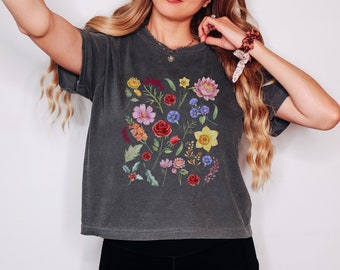 Könnte beinhalten: Ein dunkelgraues T-Shirt mit einem Blumendruck, der verschiedene Blumen in den Farben Rosa, Rot, Gelb, Blau und Grün zeigt.