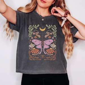 May include: A dark gray cropped t-shirt with a graphic print of a butterfly, mushrooms, flowers, and a crescent moon. The graphic is in shades of pink, orange, yellow, and green.