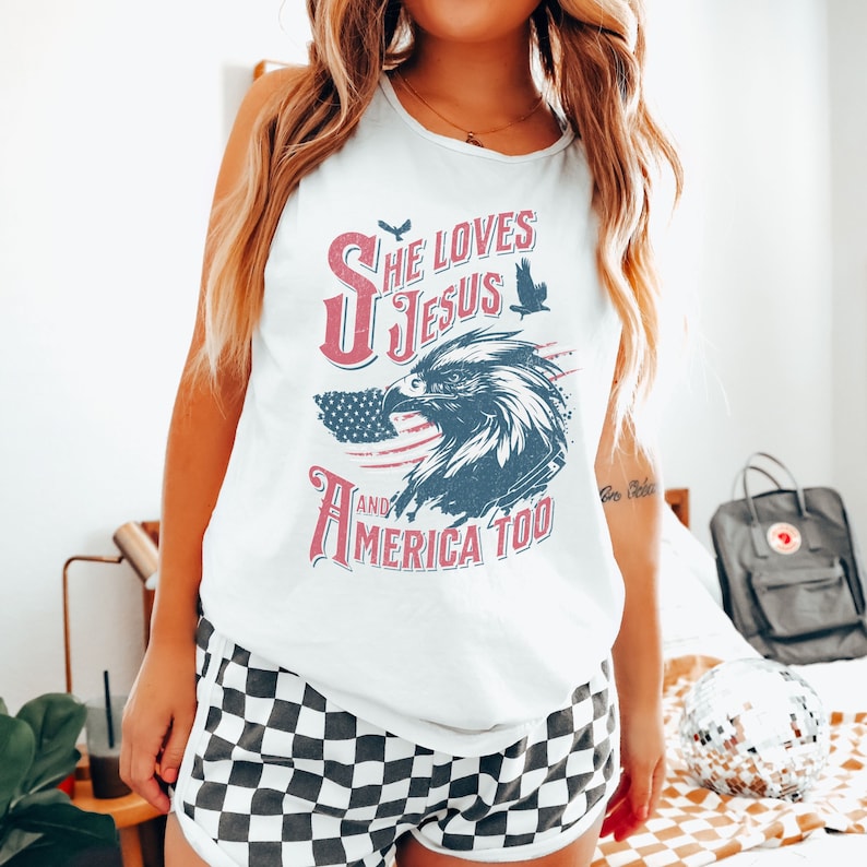 May include: White sleeveless tank top with a red, white, and blue American flag design and the text "She Loves Jesus and America Too".