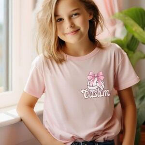 May include: A pink t-shirt with a white volleyball with a pink bow on top. The text "Custom" is printed below the volleyball.
