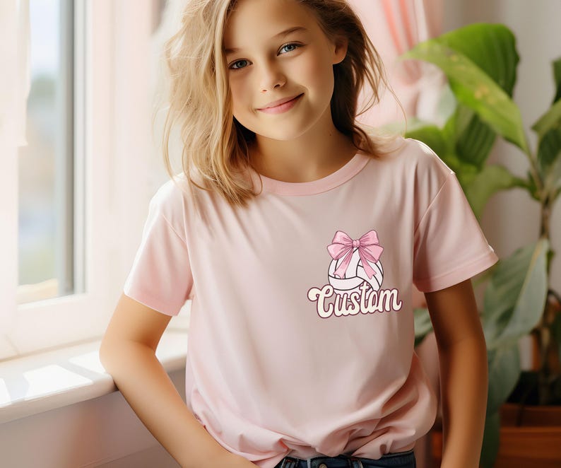 May include: A pink t-shirt with a white volleyball with a pink bow on top. The text "Custom" is printed below the volleyball.