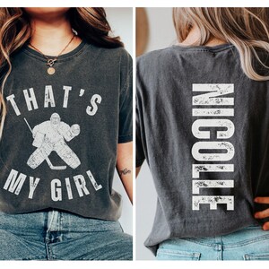 May include: A black t-shirt with a white graphic of a hockey goalie and the text "That's My Girl". The back of the shirt has the name "Nicole" printed in white.