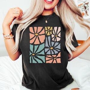 Op de afbeelding: Een zwart T-shirt met een grafisch ontwerp van kleurrijke abstracte bloemen. De bloemen zijn in een vierkant patroon gerangschikt en hebben een retro-stijl.