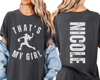 May include: A charcoal gray t-shirt featuring a distressed white graphic on the front with a silhouette of a softball player and the text "THAT'S MY GIRL". The back of the shirt displays the name "NICOLE" in large, distressed white vertical lettering.