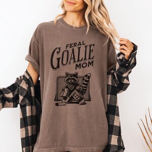 May include: A brown t-shirt with the text "Feral Goalie Mom" and a graphic of a raccoon wearing a hockey goalie mask and holding a hockey stick.