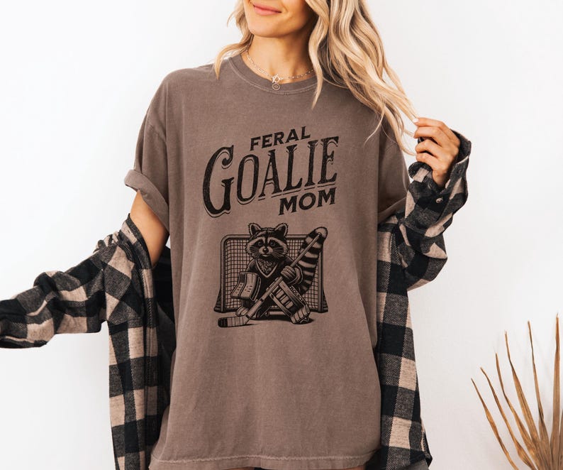 May include: A brown t-shirt with the text "Feral Goalie Mom" and a graphic of a raccoon wearing a hockey goalie mask and holding a hockey stick.