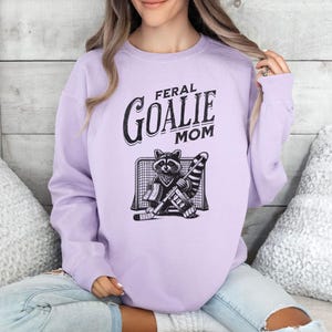 May include: A lavender sweatshirt with the text "Feral Goalie Mom" and a cartoon raccoon wearing goalie gear.