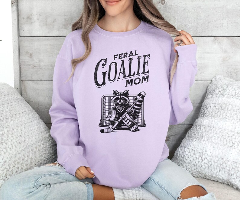 May include: A lavender sweatshirt with the text "Feral Goalie Mom" and a cartoon raccoon wearing goalie gear.