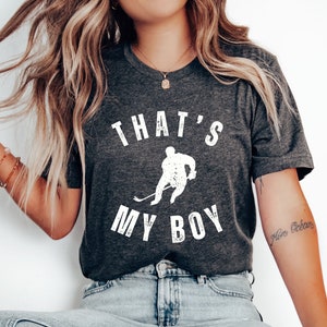 Może przedstawiać: Szary t-shirt z białym nadrukiem hokeisty i tekstem "That's My Boy".