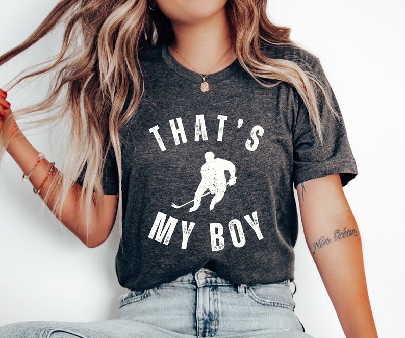 Może przedstawiać: Szary t-shirt z białym nadrukiem hokeisty i tekstem "That's My Boy".