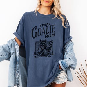 May include: A blue t-shirt with the text "Feral Goalie Mom" and a graphic of a raccoon wearing a goalie mask and holding a hockey stick.