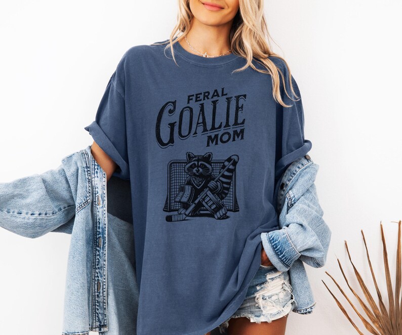 May include: A blue t-shirt with the text "Feral Goalie Mom" and a graphic of a raccoon wearing a goalie mask and holding a hockey stick.
