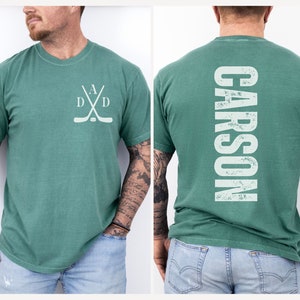 Puede incluir: Una camiseta verde con un dise&ntilde;o de palo de hockey y disco en la parte delantera y la palabra "CARSON" en la parte trasera.