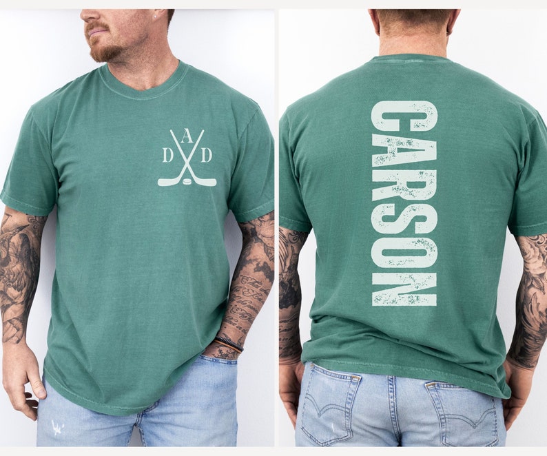 Puede incluir: Una camiseta verde con un dise&ntilde;o de palo de hockey y disco en la parte delantera y la palabra "CARSON" en la parte trasera.