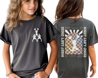 May include: A charcoal gray t-shirt featuring a white graphic of three cheerleaders in a pyramid on the front chest. The back displays a large arched checkerboard design with a jumping cheerleader and the text SORRY CAN'T CHEER BYE.