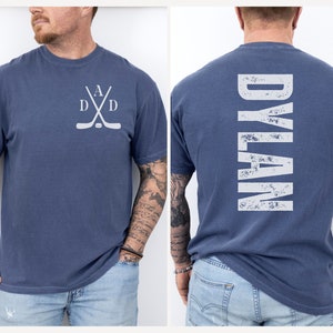 Puede incluir: Una camiseta azul con un dise&ntilde;o de palo de hockey blanco en la parte delantera y la palabra "DYLAN" en blanco en la parte posterior.