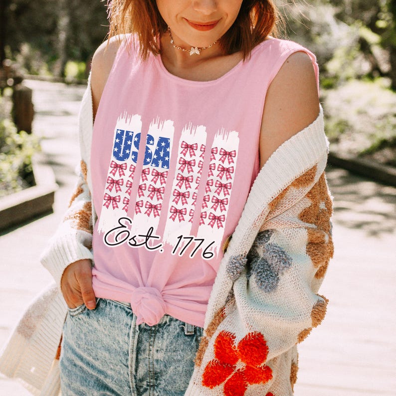 May include: A pink sleeveless top featuring a patriotic design with the word "USA" in blue and white, and red and white bows. The text "Est. 1776" is below the design. The person is wearing a white and brown patterned cardigan.