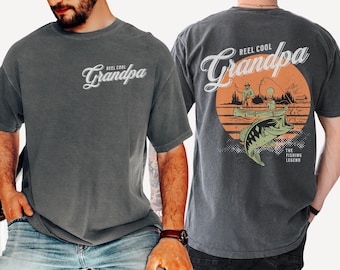 May include: A charcoal gray t-shirt featuring the text "Reel Cool Grandpa" on the front left chest. The back displays a large graphic of a person fishing in a boat on a lake with a large fish in the foreground, set against an orange sun.