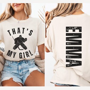 May include: A white t-shirt with the text "That's My Girl" and a hockey goalie graphic on the front. The back of the shirt has the name "EMMA" printed vertically in a distressed black font.