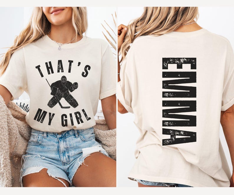 May include: A white t-shirt with the text "That's My Girl" and a hockey goalie graphic on the front. The back of the shirt has the name "EMMA" printed vertically in a distressed black font.