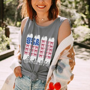May include: Grey tank top featuring a patriotic design with the word "USA" in blue and white, and red and white bows. The text "Est. 1776" is below the design. The tank top is knotted at the waist.
