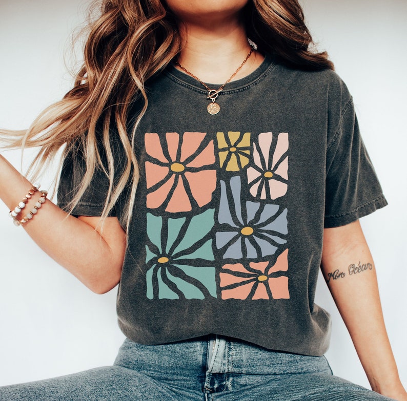 Op de afbeelding: Een donkergrijs T-shirt met een kleurrijk abstract bloemenontwerp. Het ontwerp bevat verschillende bloemen in tinten roze, blauw, groen en geel.
