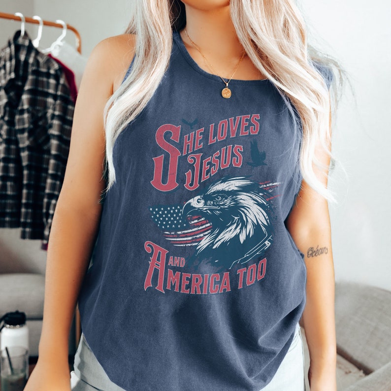 May include: A navy blue tank top with a red, white, and blue American flag design. The text "She Loves Jesus and America Too" is printed on the front of the shirt.
