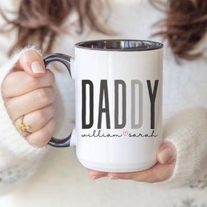 May include: White ceramic mug with a black rim and handle. The mug has the word "DADDY" printed in black and gray letters with the text "william ♥ sarah" printed below in black.