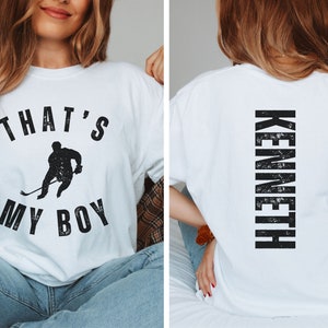 Może przedstawiać: Biała koszulka z czarnym nadrukiem hokeisty i tekstem "That's My Boy". Z tyłu koszulki znajduje się napis "Kenneth" w kolorze czarnym.