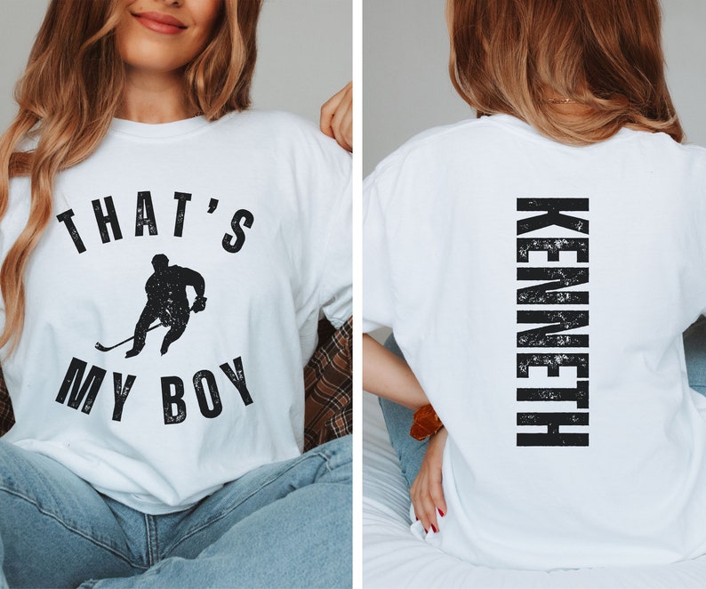 Może przedstawiać: Biała koszulka z czarnym nadrukiem hokeisty i tekstem "That's My Boy". Z tyłu koszulki znajduje się napis "Kenneth" w kolorze czarnym.