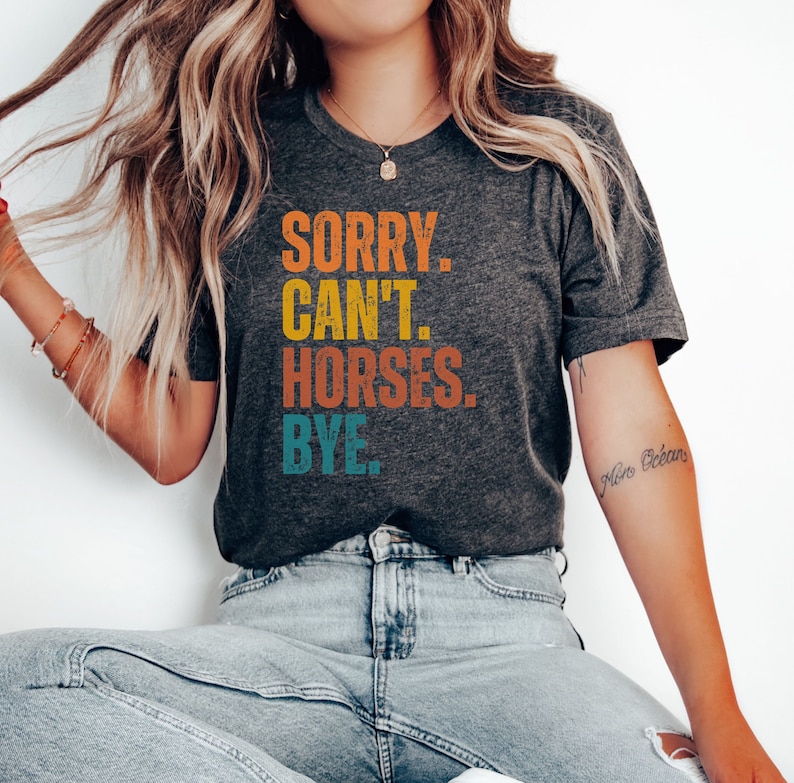 May include: A dark grey t-shirt with the text "SORRY. CAN'T. HORSES. BYE." printed in a vintage style with colorful lettering.