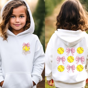 May include: White hooded sweatshirt with a softball and pink bow design on the front. The back of the sweatshirt has multiple softball and pink bow designs. The name "Kylie" is printed on the front of the sweatshirt.