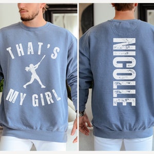 May include: A blue sweatshirt with a white graphic on the front that says "That's My Girl" with a silhouette of a woman playing baseball. The back of the sweatshirt has the name "Nicole" printed in white.