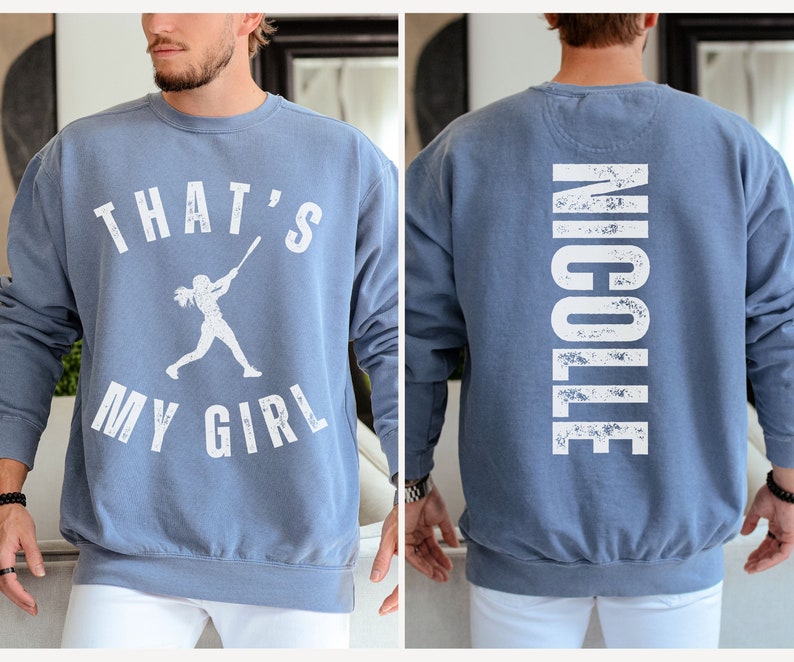 May include: A blue sweatshirt with a white graphic on the front that says "That's My Girl" with a silhouette of a woman playing baseball. The back of the sweatshirt has the name "Nicole" printed in white.