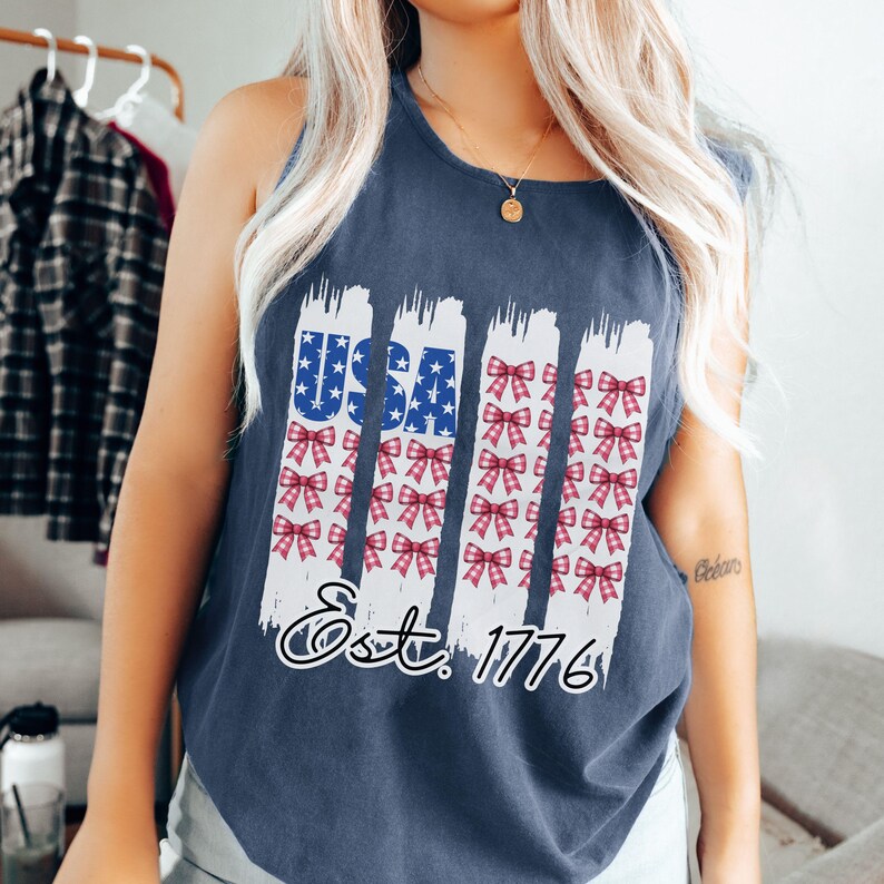 May include: Navy blue vest top with a patriotic design. The design features the word "USA" in blue with white stars, and vertical white stripes with red bows. The text "Est. 1776" is below the design.