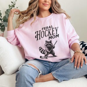 May include: A pink sweatshirt with the text "Feral Hockey Mom" in black lettering. A cartoon raccoon wearing a hockey helmet and holding a hockey stick is printed below the text.