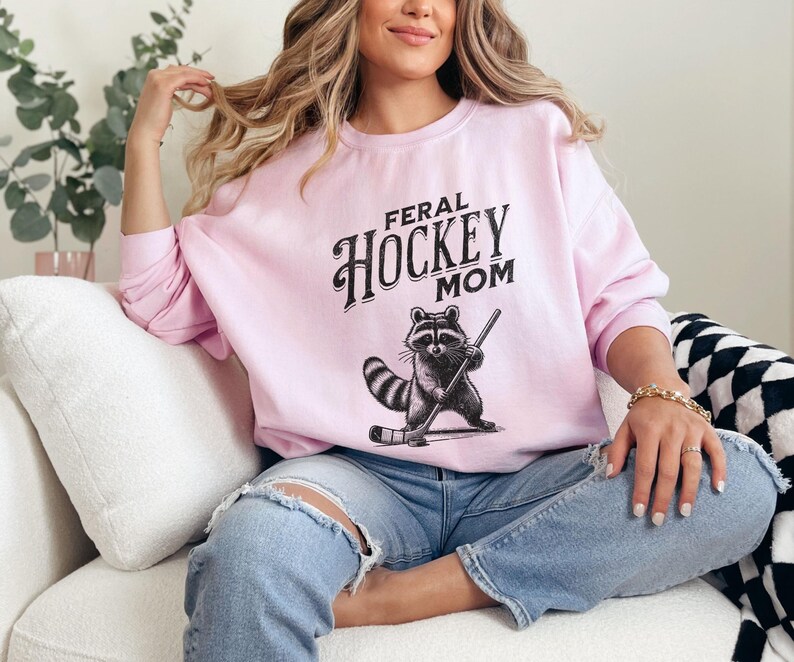May include: A pink sweatshirt with the text "Feral Hockey Mom" in black lettering. A cartoon raccoon wearing a hockey helmet and holding a hockey stick is printed below the text.