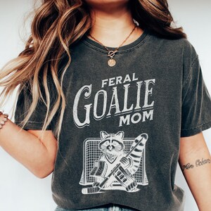 May include: A dark gray t-shirt with a white graphic of a raccoon wearing a hockey goalie uniform and the text "Feral Goalie Mom".